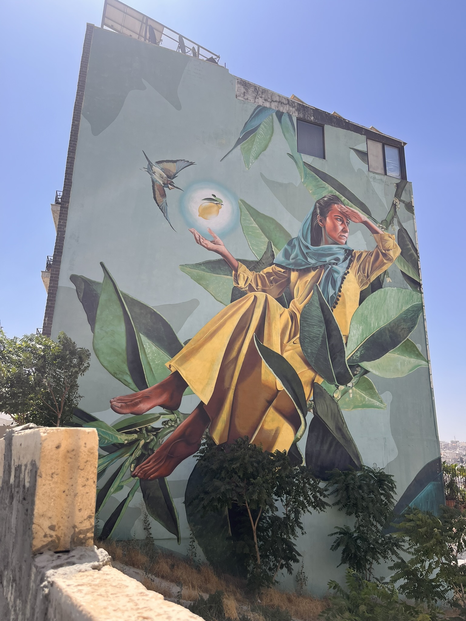 Large street mural on a building wall in Jabal Al-Weibdeh Amman — woman in yellow dress with a bird and lemon